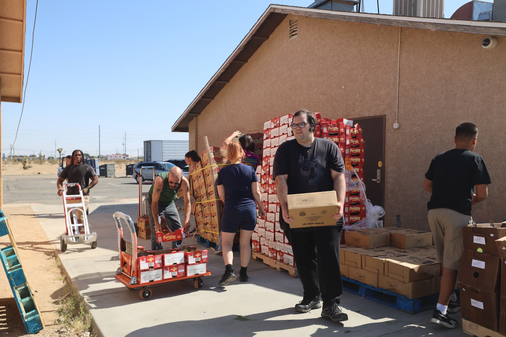 Volunteers outside a tan building moving cases of food on hand trucks and carts, with one volunteer carrying a box in the foreground.